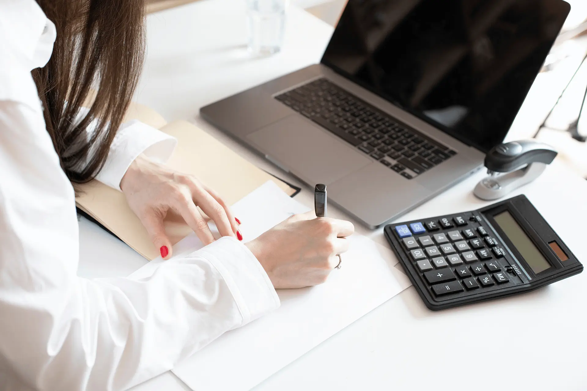 Frau auf Arbeit schreibt Eine Frau sitzt am Schreibtisch vor einem Laptop und einem Taschenrechner und schreibt etwas auf einen Zettel